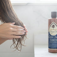 Woman applying One N Only Argan Oil Color Oasis Smoothing Conditioner to wet hair, with product bottle on bathroom counter.