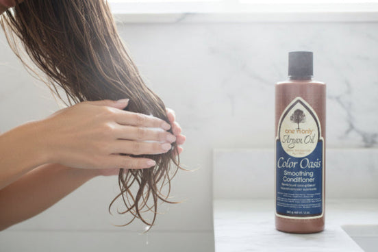 Woman applying One N Only Argan Oil Color Oasis Smoothing Conditioner to wet hair, with product bottle on bathroom counter.