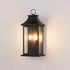 A black outdoor wall lantern light fixture with two lit candle-style bulbs, mounted on a plain off-white wall.