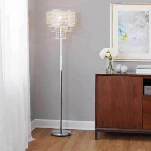 A tall crystal chandelier floor lamp with a silver metal stand and a clear faceted crystal shade, shown lit in a modern room setting next to a wooden cabinet.