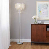 A tall crystal chandelier floor lamp with a silver metal stand and a clear faceted crystal shade, shown lit in a modern room setting next to a wooden cabinet.