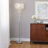 A tall crystal chandelier floor lamp with a silver metal stand and a clear faceted crystal shade, shown lit in a modern room setting next to a wooden cabinet.
