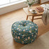 Floral patterned ottoman on a wooden floor with a table and cup in the background