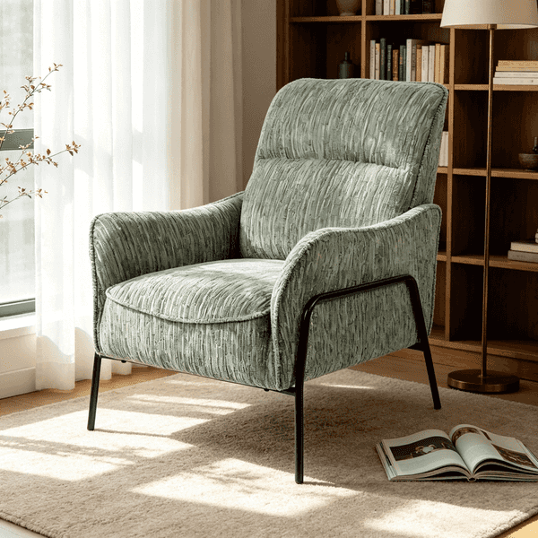 A modern upholstered armchair in textured sage green fabric with black metal legs, placed on a light rug in a sunlit room with a bookshelf and floor lamp in the background.