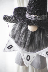 A close-up of a decorative gnome figure wearing a black glitter witch hat with a striped band, a long gray faux-fur beard, and a white pennant banner that reads BOO.