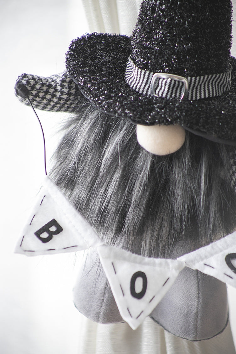 A close-up of a decorative gnome figure wearing a black glitter witch hat with a striped band, a long gray faux-fur beard, and a white pennant banner that reads BOO.