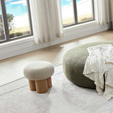 Two round ottomans, one with wooden legs and the other with a blanket draped over it, in a room with large windows.
