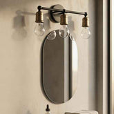 A three-light wall sconce with clear bulb-shaped glass shades and antique brass fittings mounted above an oval wall mirror on a textured beige wall.