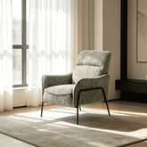A modern upholstered accent chair in textured sage green fabric with black metal legs, positioned on a neutral rug in a brightly lit room with sheer curtains and natural sunlight.