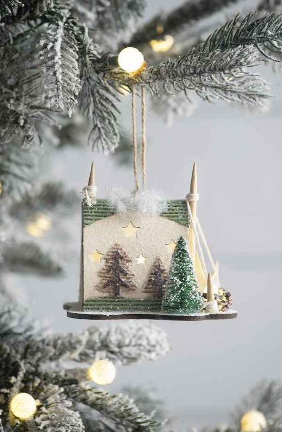 A small hanging Christmas ornament shaped like a miniature house with snowy trees, stars, and a snowy roof, suspended by twine on a flocked Christmas tree branch with warm blurred lights in the background.