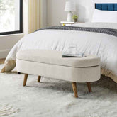 Beige ottoman with wooden legs in a bedroom setting.