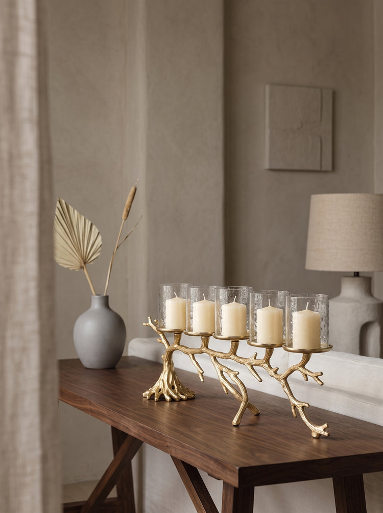 A gold branch-shaped candle holder with five clear glass candle holders and white pillar candles, styled on a dark wood console table in a warm, neutral-toned living room.