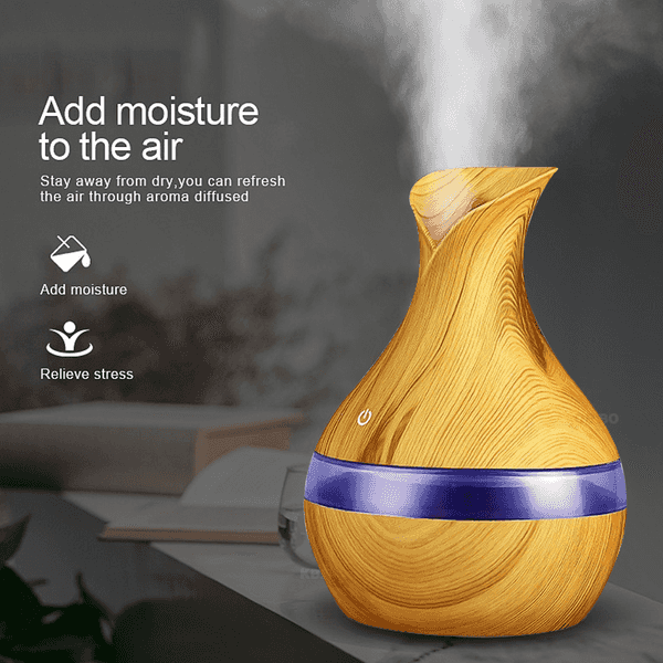 A wooden-look essential oil diffuser with a purple band, emitting steam, shown on a tabletop with promotional text and blurred background elements.