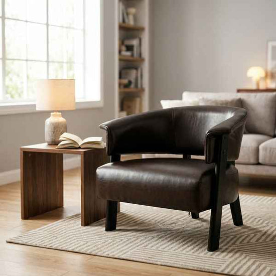 A dark brown leather club chair with black legs, styled in a cozy living room next to a wooden side table with a lamp and book.