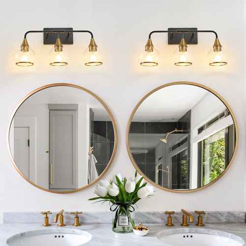 Two round wall mirrors with gold frames mounted above a double bathroom vanity with gold faucets and a lit three-light wall sconce fixture on each side.