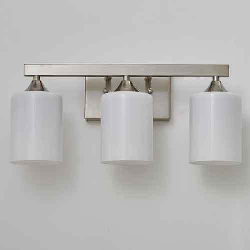 A three-light wall-mounted vanity light fixture with cylindrical white shades and a brushed nickel metal bar and backplate, shown against a plain light gray wall.