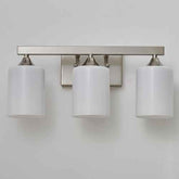 A three-light wall-mounted vanity light fixture with cylindrical white shades and a brushed nickel metal bar and backplate, shown against a plain light gray wall.