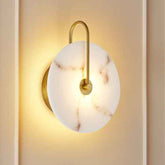A modern wall sconce with a round white marble-look glass shade and a curved gold metal arm, shown illuminated against a warm beige wall.