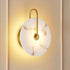 A modern wall sconce with a round white marble-look glass shade and a curved gold metal arm, shown illuminated against a warm beige wall.