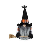 A Halloween witch gnome decoration with a tall black pointed hat topped by a small bat figure, an orange, black, and white plaid band, a gray faux-fur beard, black hands, and a broom on a wooden handle.