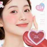 A close-up of a smiling woman with pink rosy cheeks holding a heart-shaped compact of the same blush shade. The compact has a pink border and the words &