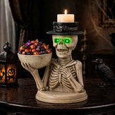 Halloween skeleton candle holder figurine holding a bowl of candy, with a lit white candle on its hat and glowing green eyes, displayed on a dark wooden table with a lantern and raven in a spooky room.