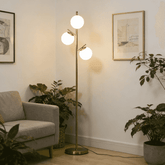 A modern floor lamp with a slim brass-colored metal pole and three spherical white glass shades, illuminated in a cozy living room corner next to a gray armchair and potted plants.