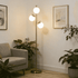 A modern floor lamp with a slim brass-colored metal pole and three spherical white glass shades, illuminated in a cozy living room corner next to a gray armchair and potted plants.