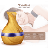 A teardrop-shaped wooden-look essential oil or humidifier diffuser with a blue band, shown in front of a sleeping woman on a white background. Text reads &