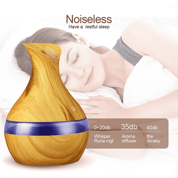 A teardrop-shaped wooden-look essential oil or humidifier diffuser with a blue band, shown in front of a sleeping woman on a white background. Text reads &