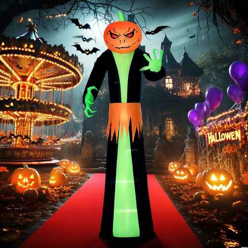 A Halloween-themed inflatable figure stands on a red carpet in a spooky village scene. The figure has a glowing green neck and torso, black arms and legs, and an orange pumpkin head with a stitched angry expression.