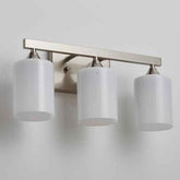 A three-light bathroom vanity wall sconce with a brushed nickel or brass horizontal bar and three white cylindrical frosted glass shades, shown mounted on a light gray wall.