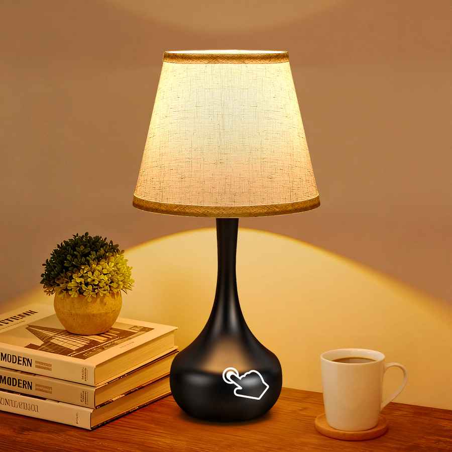 Black table lamp with a white shade on a wooden surface next to books and a cup.