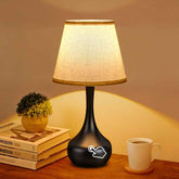 Black table lamp with a white shade on a wooden surface next to books and a cup.