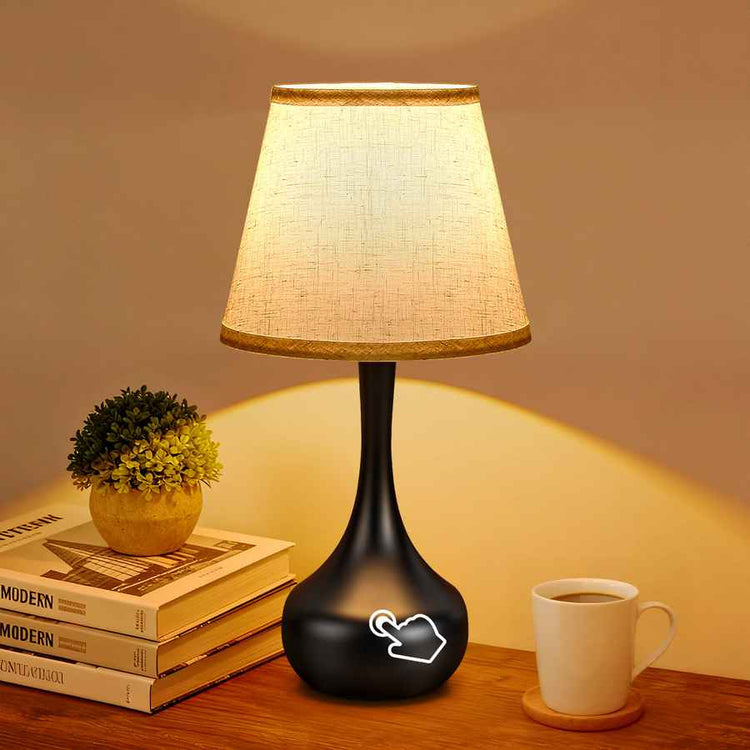 Black table lamp with a white shade on a wooden surface next to books and a cup.
