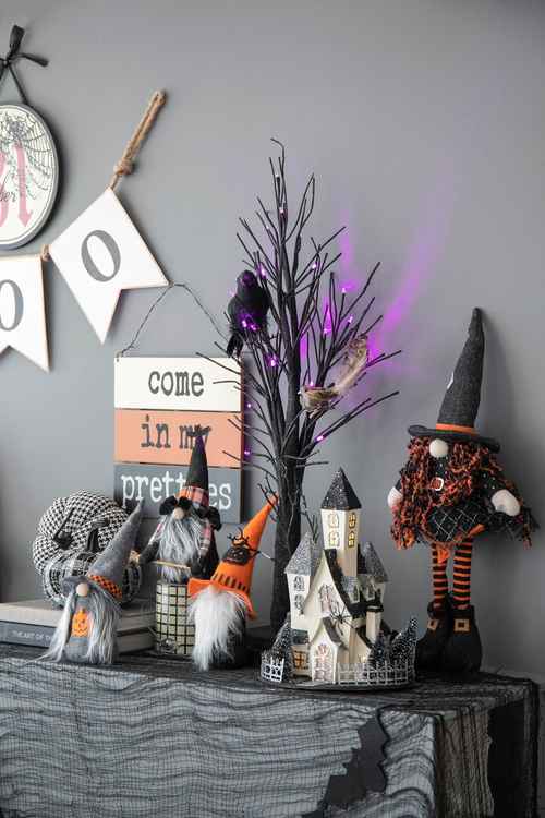 A styled Halloween tabletop display against a gray wall, featuring a witch doll, small gnomes, a haunted house, bare black branches with purple lights, and a hanging sign that reads &