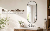 An oval bathroom mirror with a thin black frame hangs on a beige wall above a white and wood vanity. The mirror reflects a bright window, and the scene includes the text &