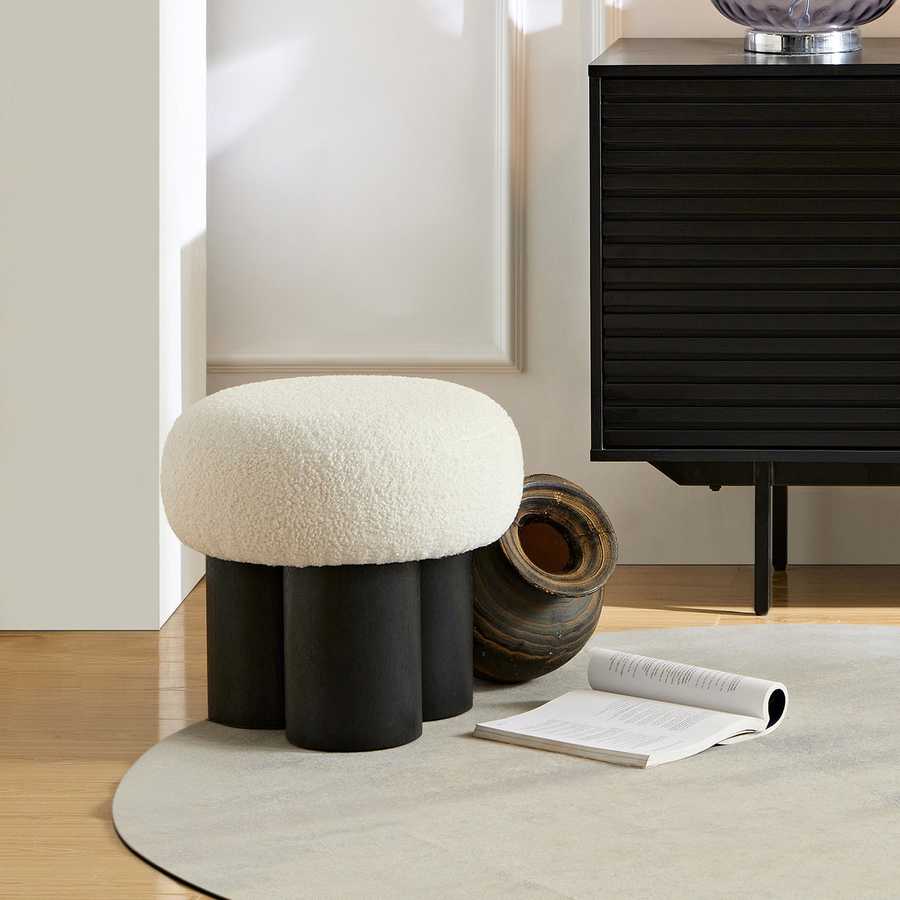 Round white cushioned stool with black cylindrical legs in a room setting.