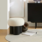 Round white cushioned stool with black cylindrical legs in a room setting.