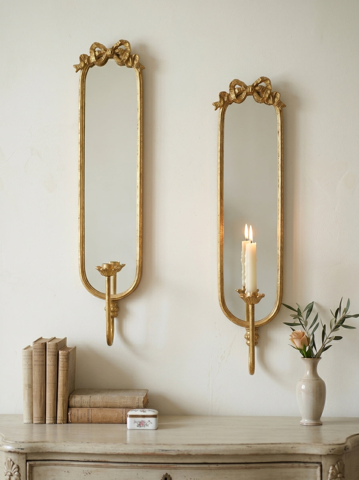Two ornate gold wall mirrors with bow-shaped tops are mounted on a cream wall above a distressed console table. One mirror is empty, and the other holds a lit white candle.