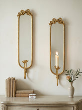 Two ornate gold wall mirrors with bow-shaped tops are mounted on a cream wall above a distressed console table. One mirror is empty, and the other holds a lit white candle.