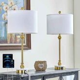 Two matching table lamps with white drum shades and brushed gold bases are displayed on a console table in a styled interior setting. The lamps have round white marble-like feet with gold trim and a small gold finial on top.