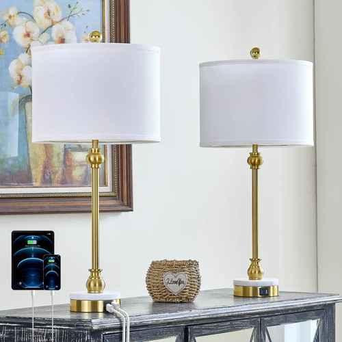 Two matching table lamps with white drum shades and brushed gold bases are displayed on a console table in a styled interior setting. The lamps have round white marble-like feet with gold trim and a small gold finial on top.