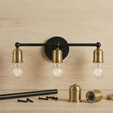 Three-light wall sconce with a black horizontal bar and round backplate, paired with brass-toned socket covers and clear Edison-style bulbs, shown mounted against a light wood wall with mounting hardware below.