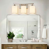 A modern three-light bathroom vanity fixture with cylindrical white shades and a brushed metal frame, mounted above a large rectangular mirror on a white wall.