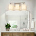A modern three-light bathroom vanity fixture with cylindrical white shades and a brushed metal frame, mounted above a large rectangular mirror on a white wall.