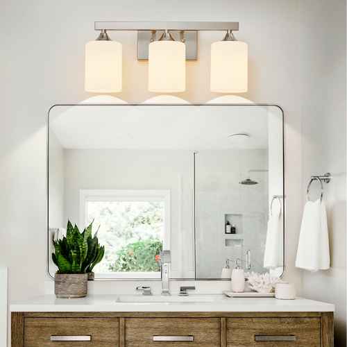 A modern three-light bathroom vanity fixture with cylindrical white shades and a brushed metal frame, mounted above a large rectangular mirror on a white wall.