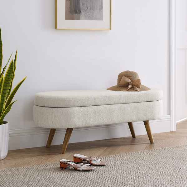 Beige storage bench with a hat on top in a room with a plant and shoes.