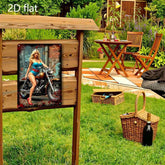 A rectangular metal sign or wall art panel mounted on a wooden display stand in a garden setting. The panel features a vintage-style pin-up illustration of a woman sitting on a motorcycle, wearing a white hat, blue top, and red skirt with red high heels.