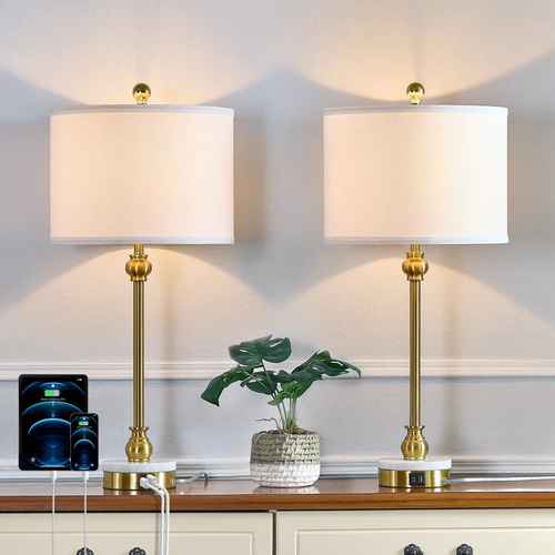 Two matching table lamps with white drum shades and gold-finished bases are shown on a wooden console against a light gray wall. A small potted green plant sits between them, and a smartphone charging dock is visible on the left.